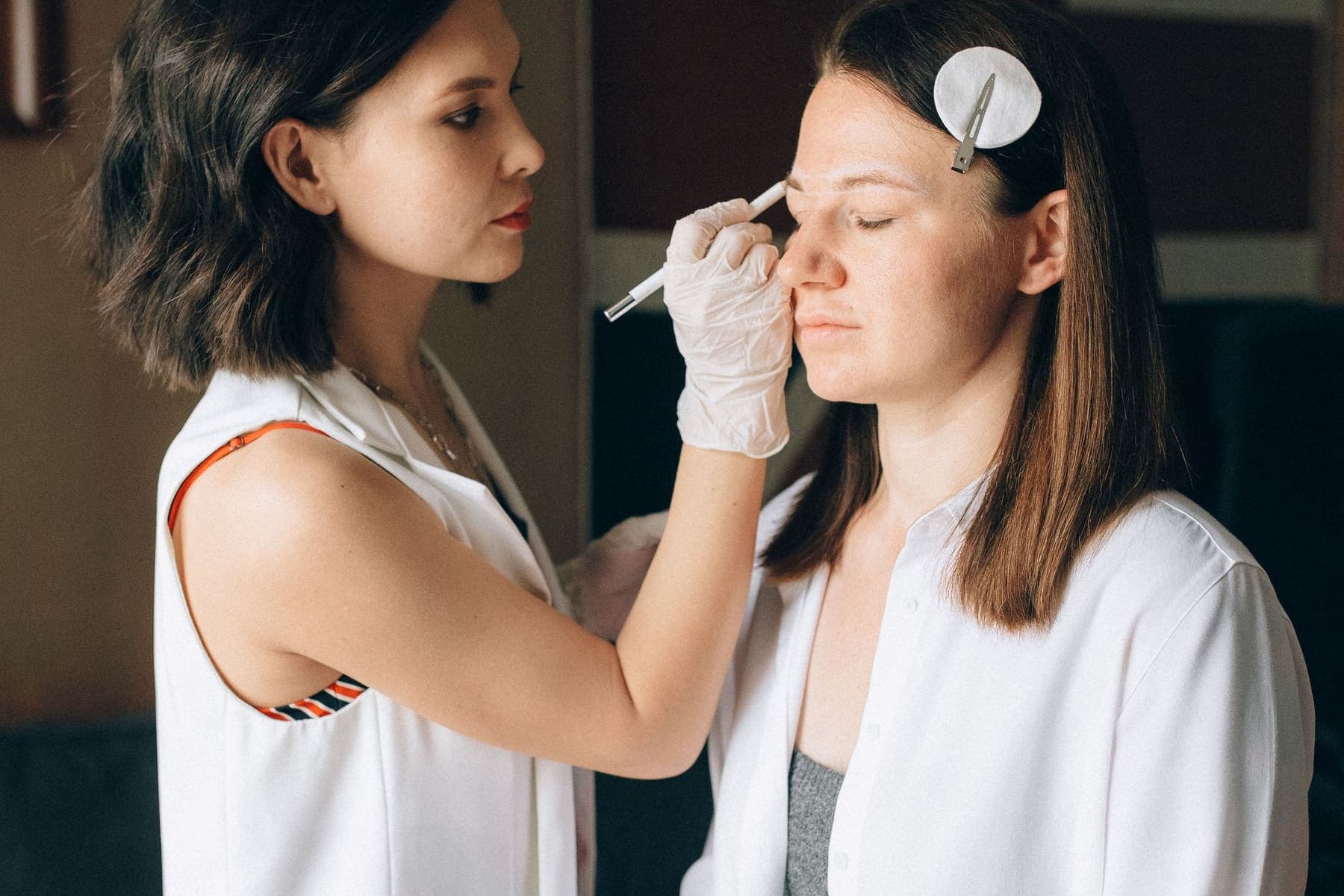 Una diseñadora de cejas con guantes marcando la ceja de una clienta sentada, con el pelo recogido con un pinche.