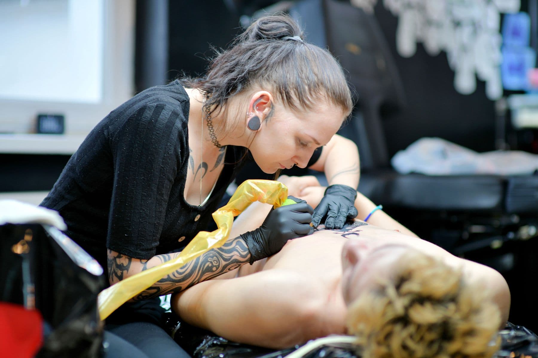 Una tatuadora con guantes negros tatuando la espalda de un cliente recostado en la camilla del estudio.